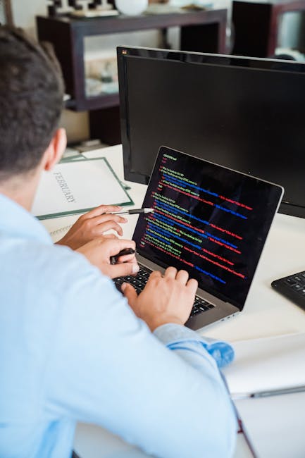 Close-up view of a computer laptop with visible screen displaying editing code, placed on a wooden desk alongside a white smartphone with a black screen. The workspace appears tidy and well-lit, highl